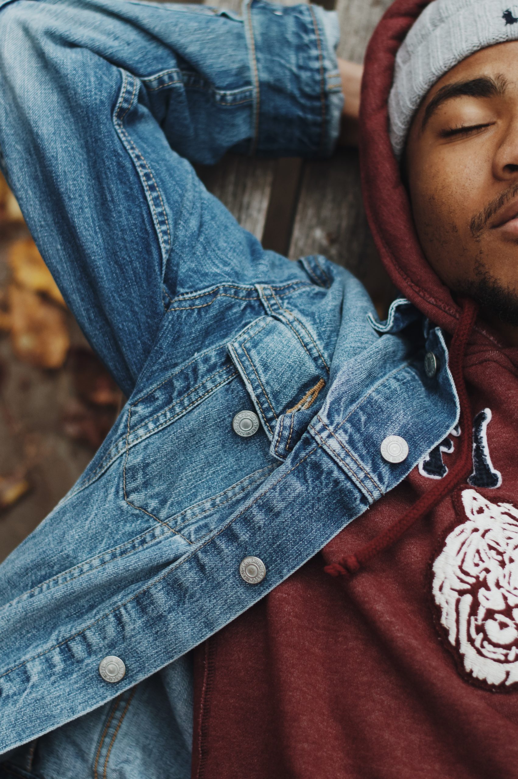 A black man wearing a red hoodie and jean jacket lying down on a park bench. Fallen leaves are in the background out of focus.