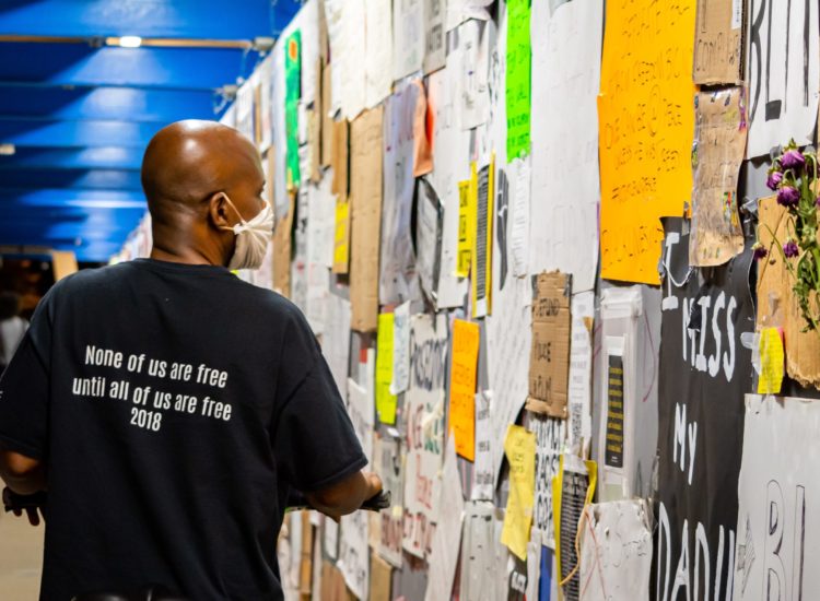 A Black man is looking at a wall full of posters wearing a shirt that reads "None of us are free until all of us are free 2018" on the back.