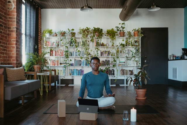 a black man in a blue shirt and white pants sitting on the ground in a meditative position. In the background there is a large bookcase full of books and many plants.