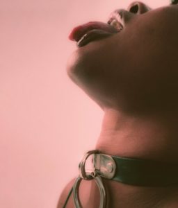a close up of a Black woman wearing a leather collar and o-ring with her tongue sticking out
